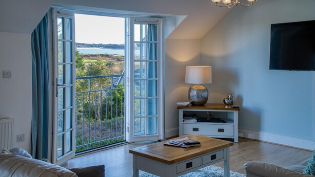 A living room with a large sliding glass door leading to a balcony, a wooden coffee table, a white TV stand, and a lamp. - Caldey View Cottage - 1 Bedroom - Penally (Tenby)