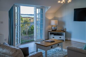 A living room with a large sliding glass door leading to a balcony, a wooden coffee table, a white TV stand, and a lamp. - Caldey View Cottage - 1 Bedroom - Penally (Tenby)