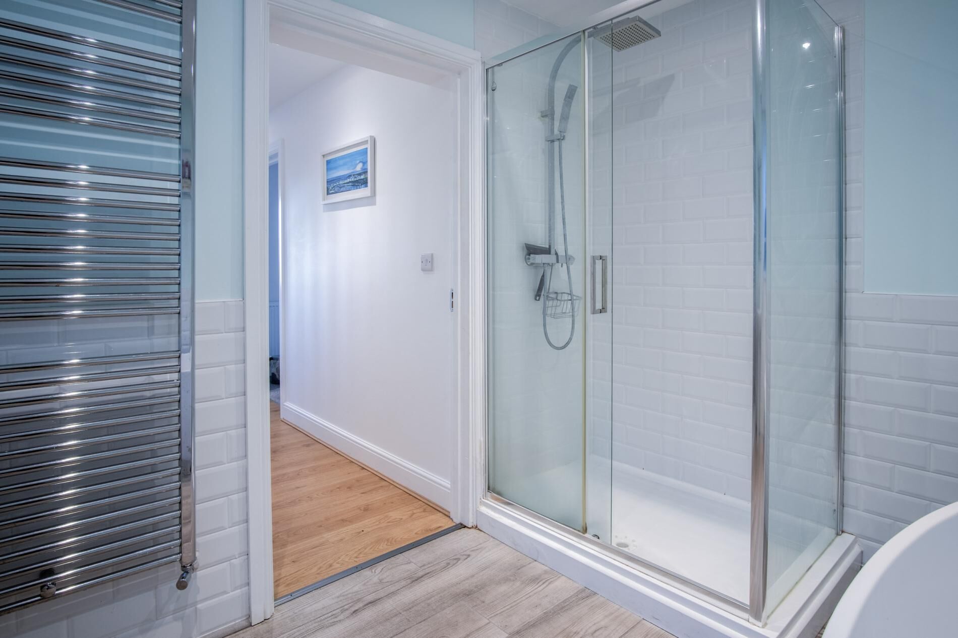 A modern bathroom with a glass-enclosed shower, white tiled walls, and a wooden floor.