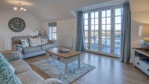 A spacious living room with a grey sofa, a coffee table, and a large window with curtains. - Caldey View Cottage - 1 Bedroom - Penally (Tenby)