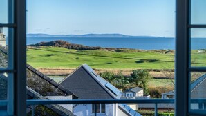 A coastal view from a window, featuring a grassy landscape, a body of water, and distant mountains. - Caldey View Cottage - 1 Bedroom - Penally (Tenby)