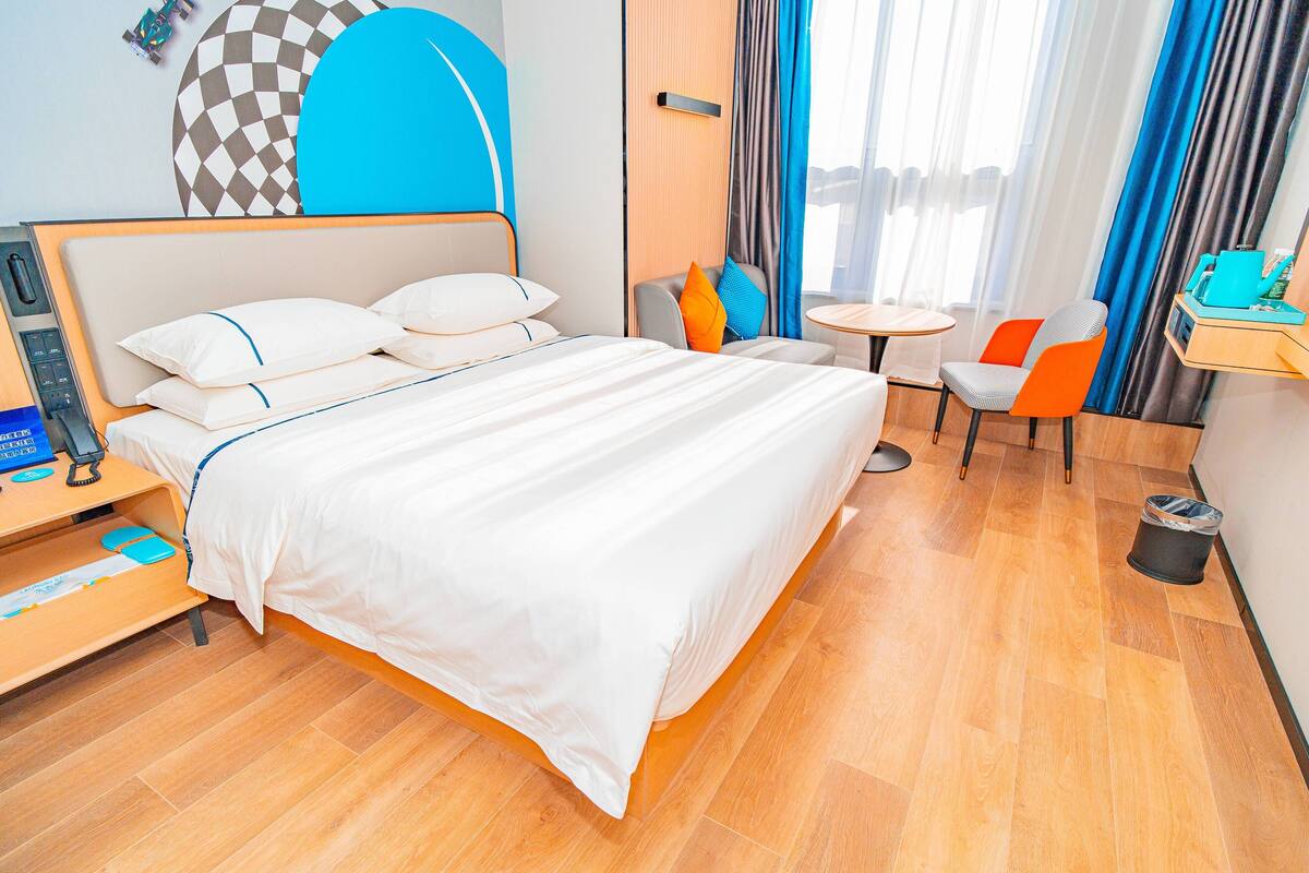 A hotel room with a bed, a desk with a phone, a chair, a small table, and a trash bin.