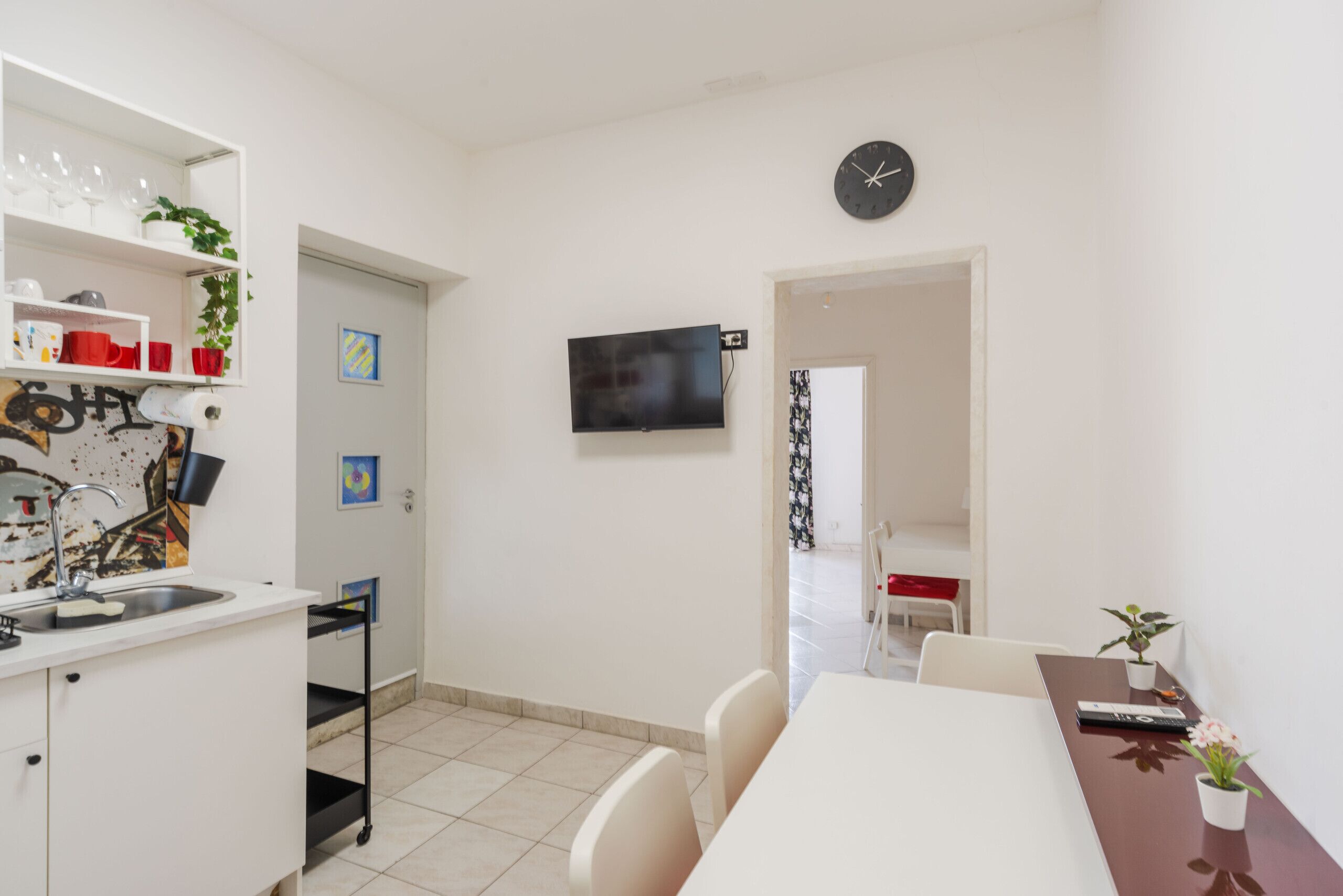 A modern kitchen with white cabinets, a sink, and a dining area with a table and chairs. A clock is mounted on the wall, and a TV is visible in the background.