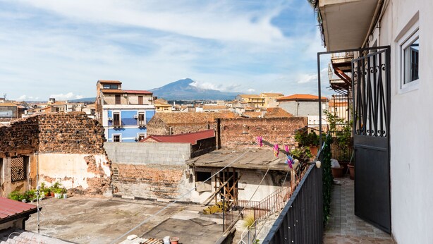 Una azotea con vistas a una montaña, una mezcla de edificios antiguos y modernos, y un balcón con plantas en macetas.