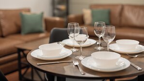 Tavolo da pranzo apparecchiato con piatti e ciotole bianche, calici da vino, e un divano in pelle marrone sullo sfondo.