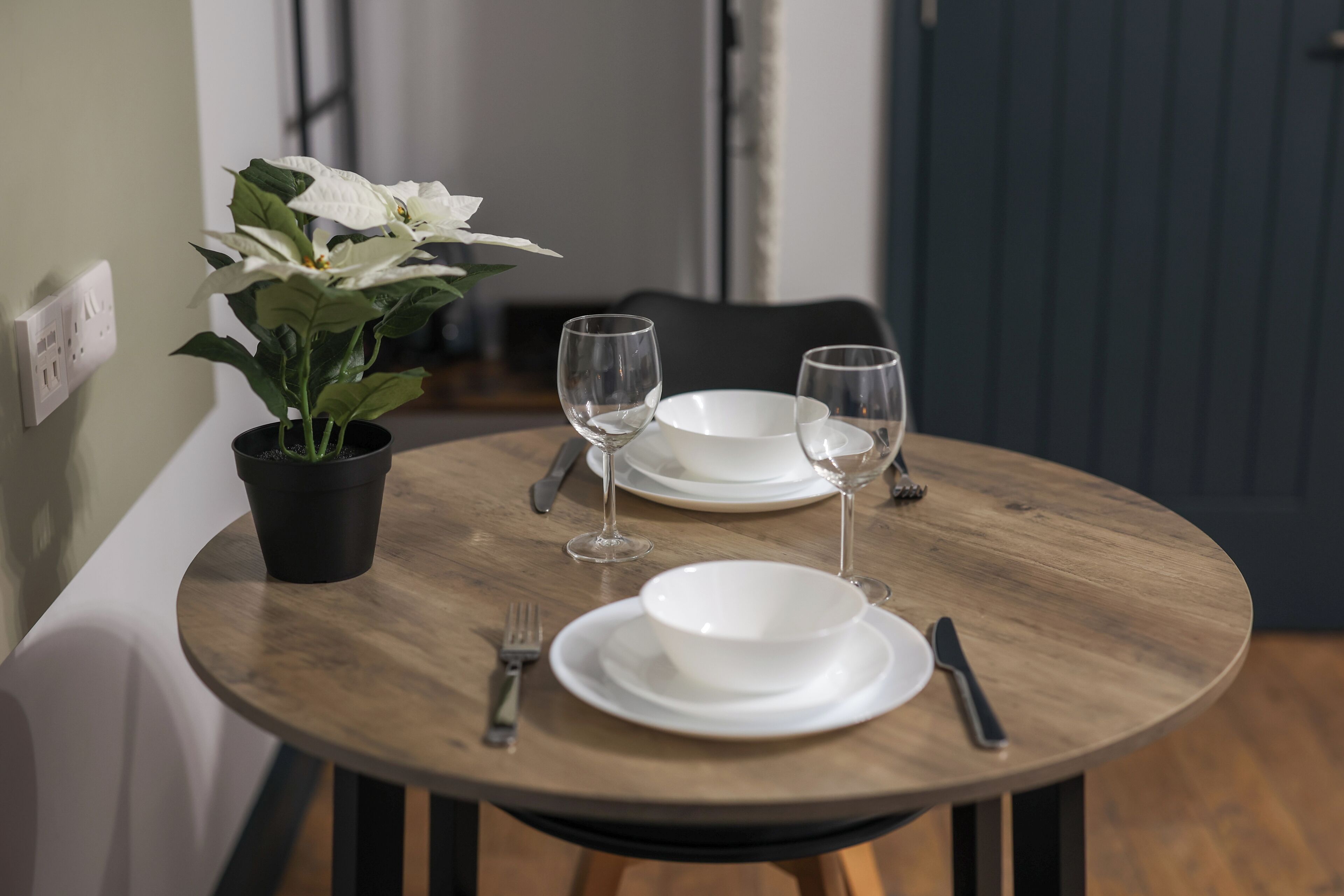 Una mesa de madera redonda con platos blancos, copas de vino y cubiertos. Hay una planta en maceta sobre la mesa.