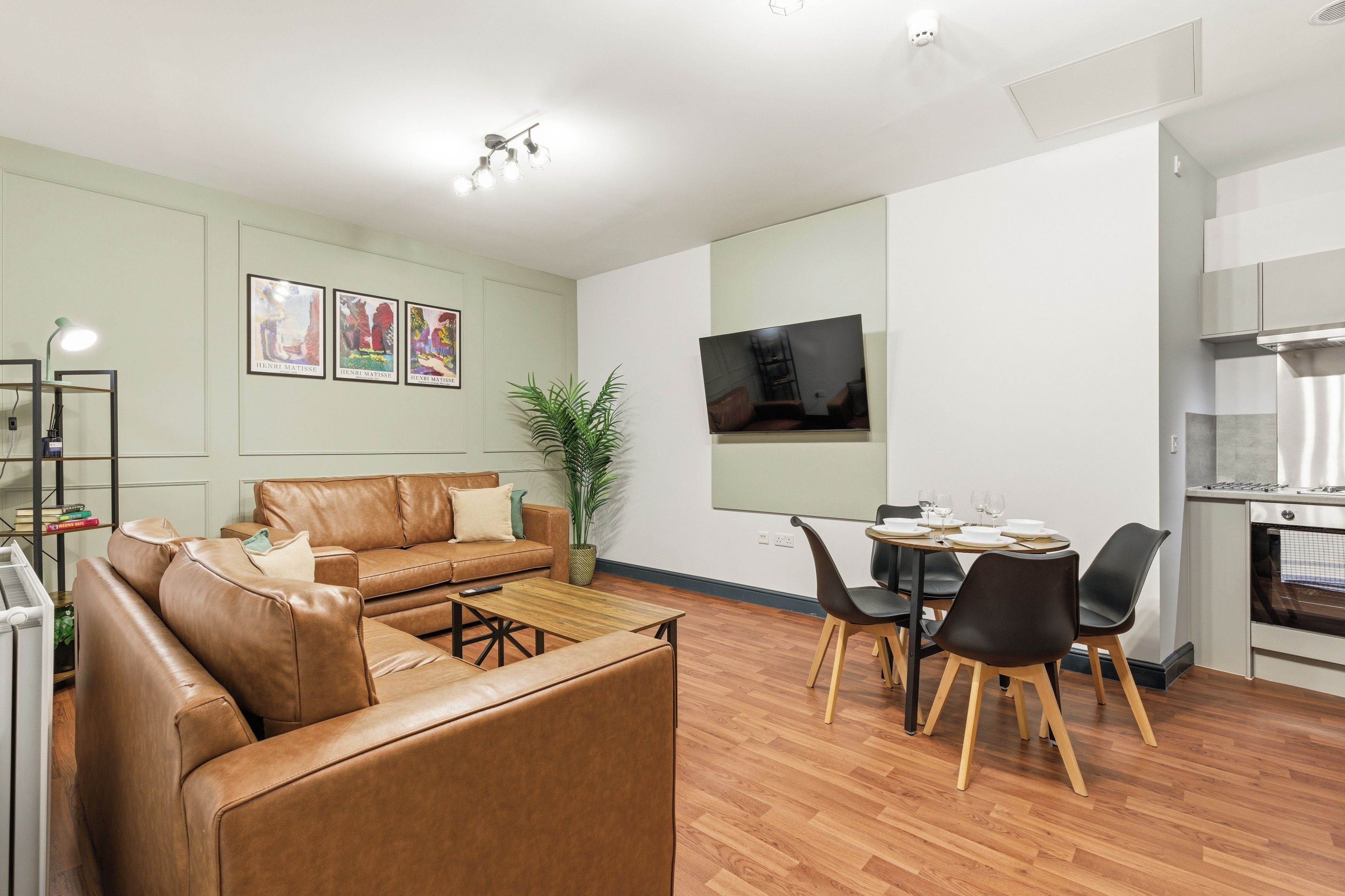 Una sala moderna con un sofá de cuero marrón, una mesa de centro, un comedor con sillas, un televisor montado en la pared y una cocina con estufa.