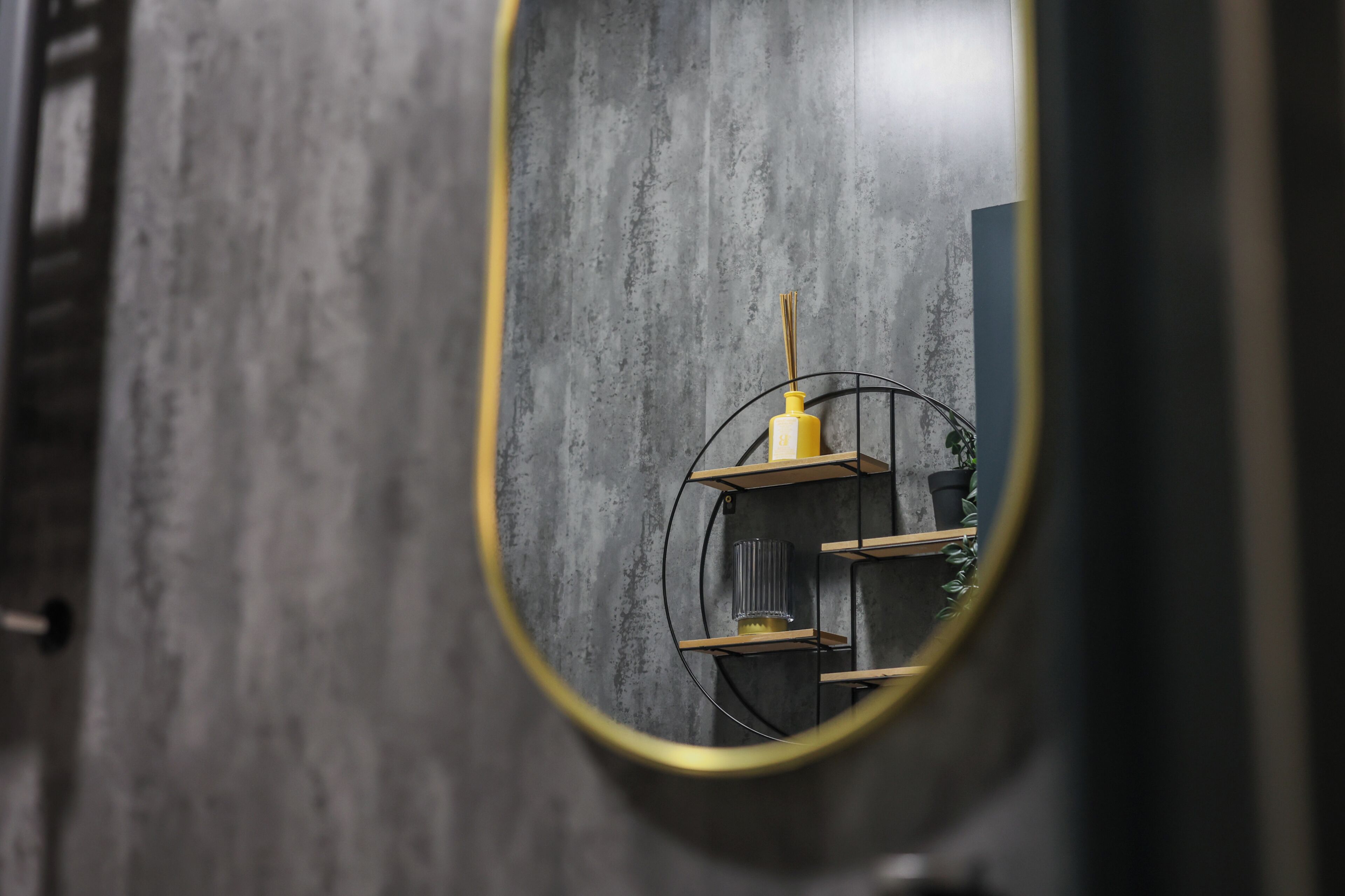 A circular mirror reflecting a modern interior with a concrete wall, a small shelf with a yellow bottle, and a potted plant.