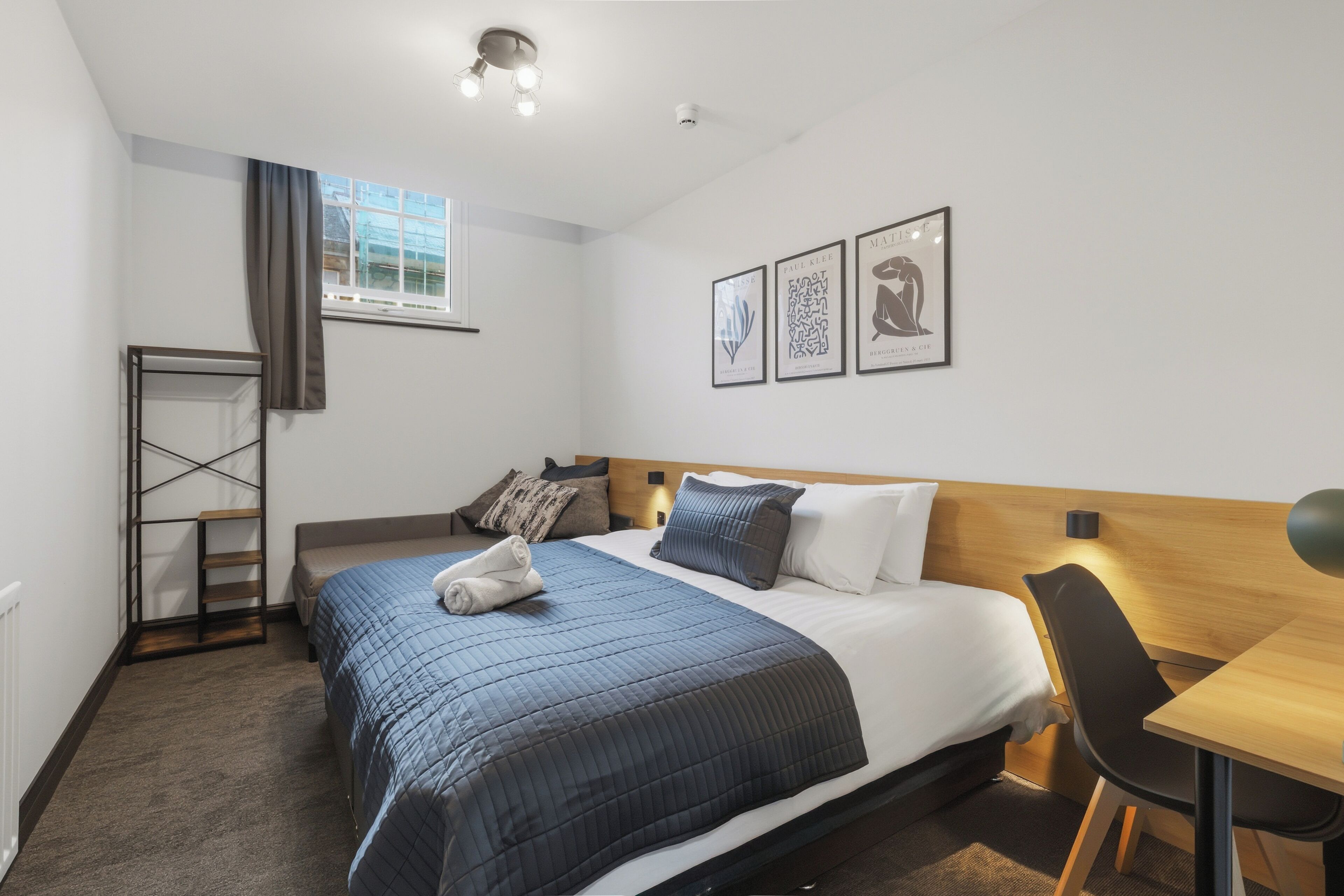 A hotel room with a bed, a desk, a chair, a window, and wall art.