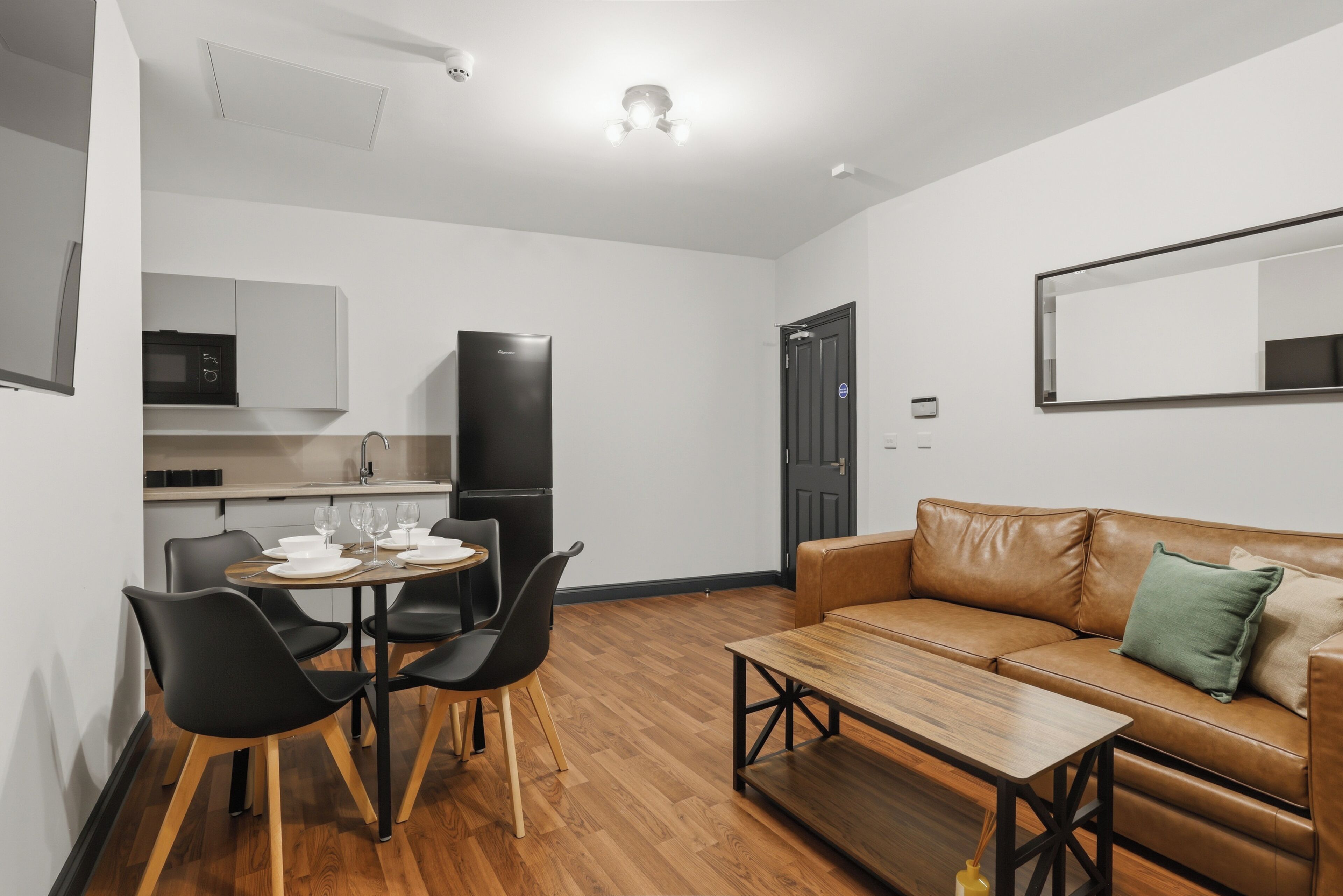 A compact living space with a dining area, a sofa, a coffee table, a kitchen with a microwave and refrigerator, and a door with a lock.