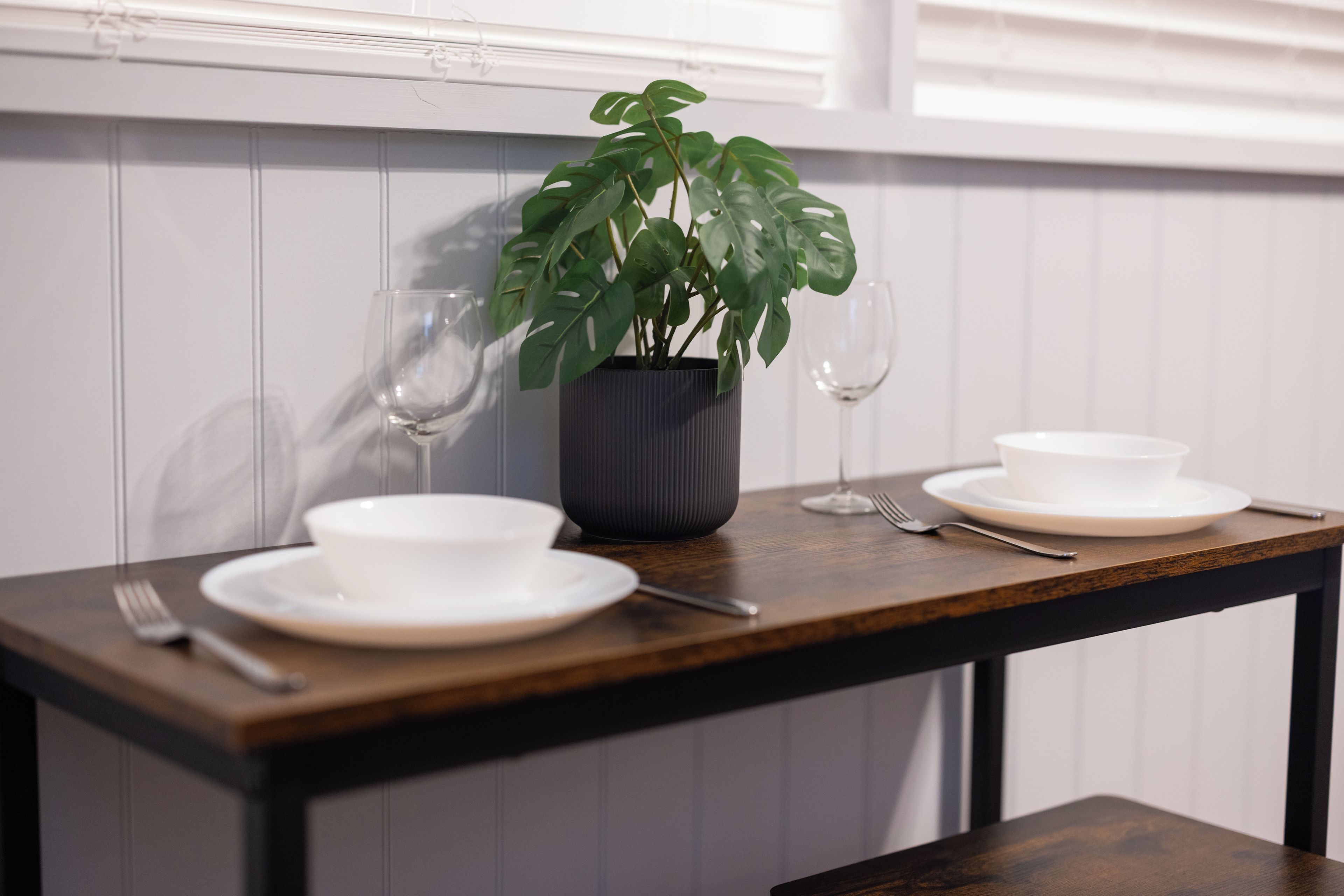 Una mesa de comedor con una planta en maceta, dos copas de vino y un juego de vajilla blanca.