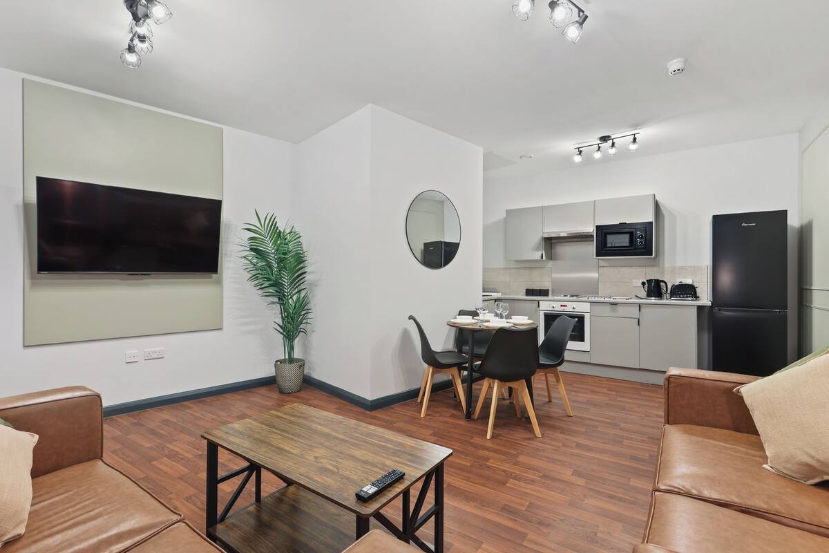 Una sala de estar moderna con un televisor de pantalla plana, una mesa de centro de madera y sofás de cuero. Hay un comedor con mesa y sillas, y una cocina con microondas y refrigerador.