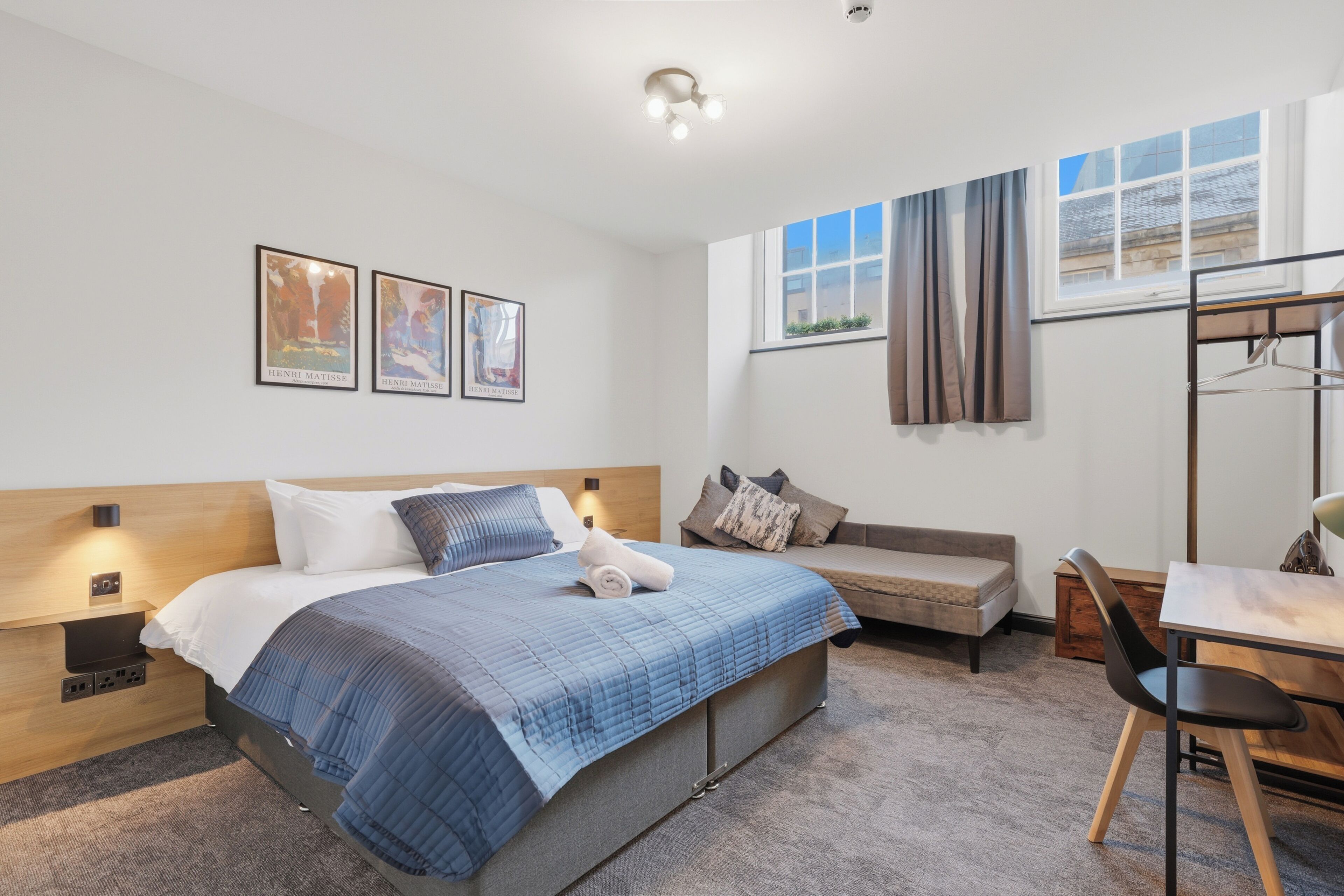 A modern hotel room with a bed, a desk, a chair, a bench, and artwork on the wall.
