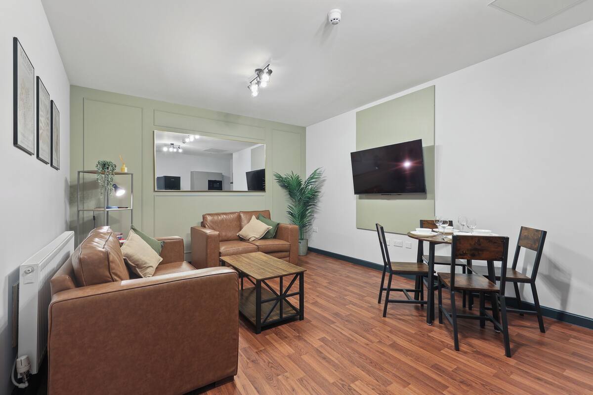 Una sala de estar moderna con un sofá de cuero marrón, una mesa de centro de madera, un área de comedor con una pequeña mesa y sillas, un espejo grande y un televisor de pantalla plana montado en la pared.