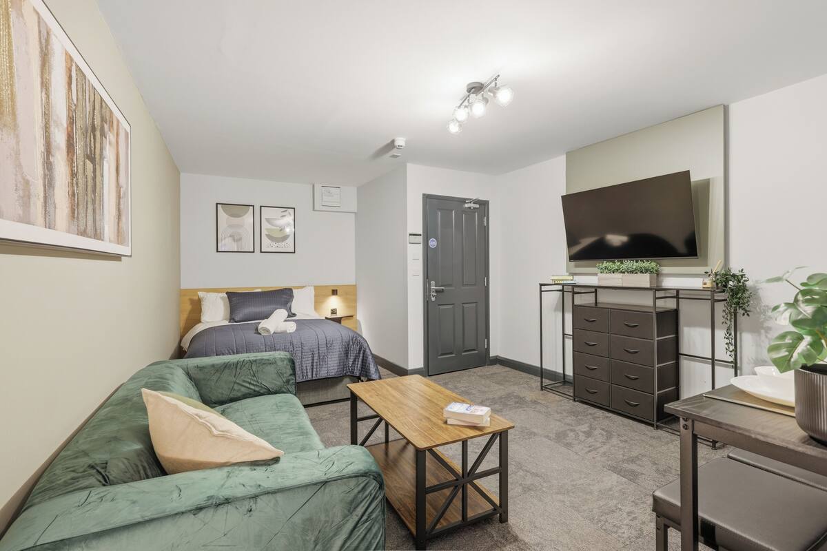 Una habitación moderna y bien iluminada con cama, sofá verde, mesa de centro de madera, cómoda y televisor de pantalla plana montado en la pared.