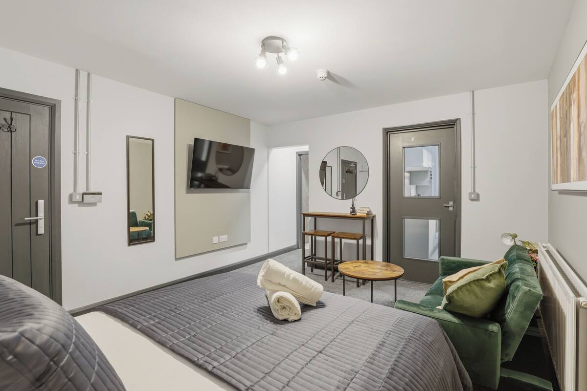 Habitación de hotel moderna con una cama, un sofá verde, una mesita redonda, un espejo y un televisor en la pared.