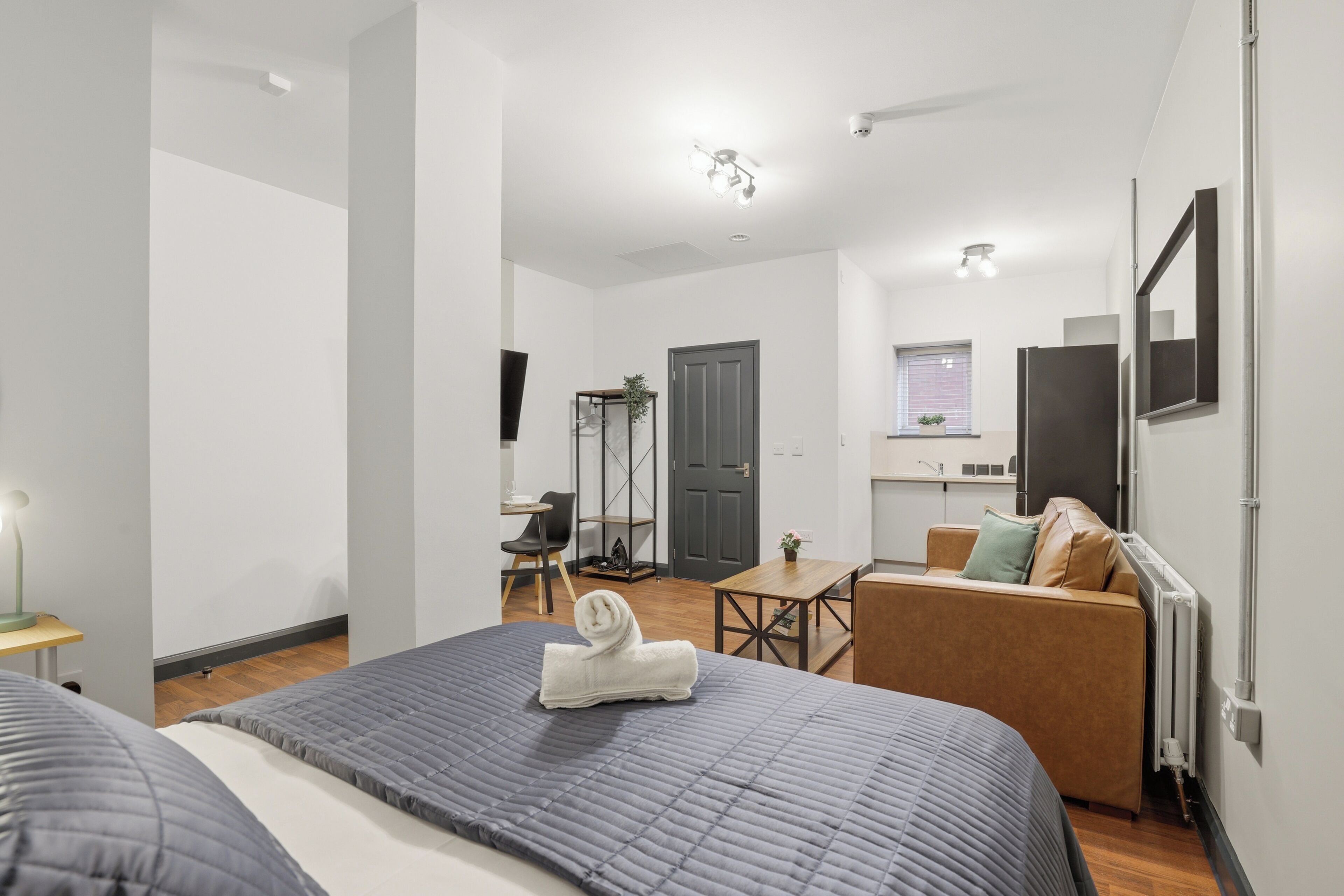 A modern hotel room with a large bed, a small desk, a sofa, and a kitchenette.
