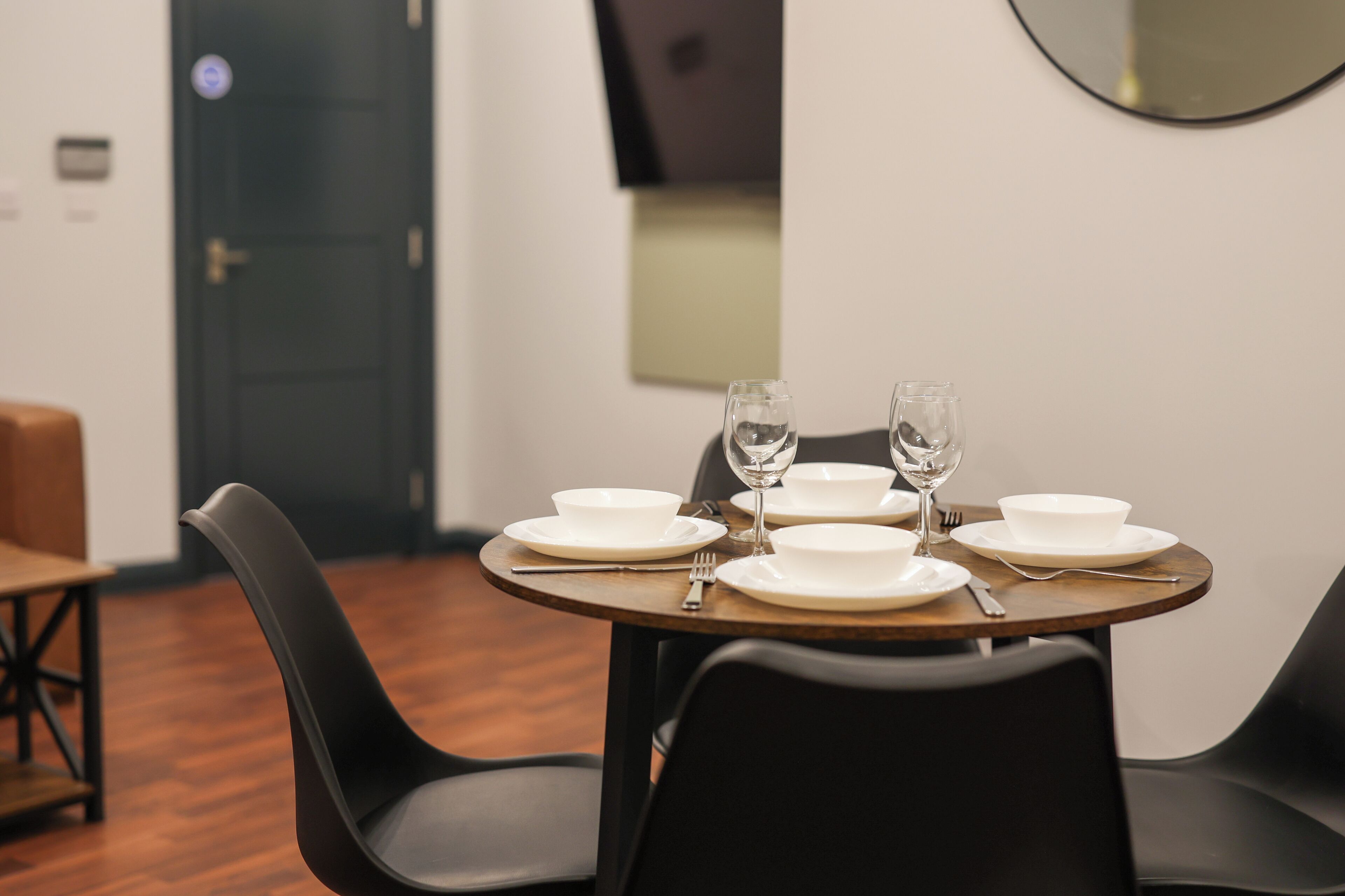 A dining table set with white plates, bowls, and wine glasses, surrounded by black chairs in a room with wooden flooring and a dark door in the background.
