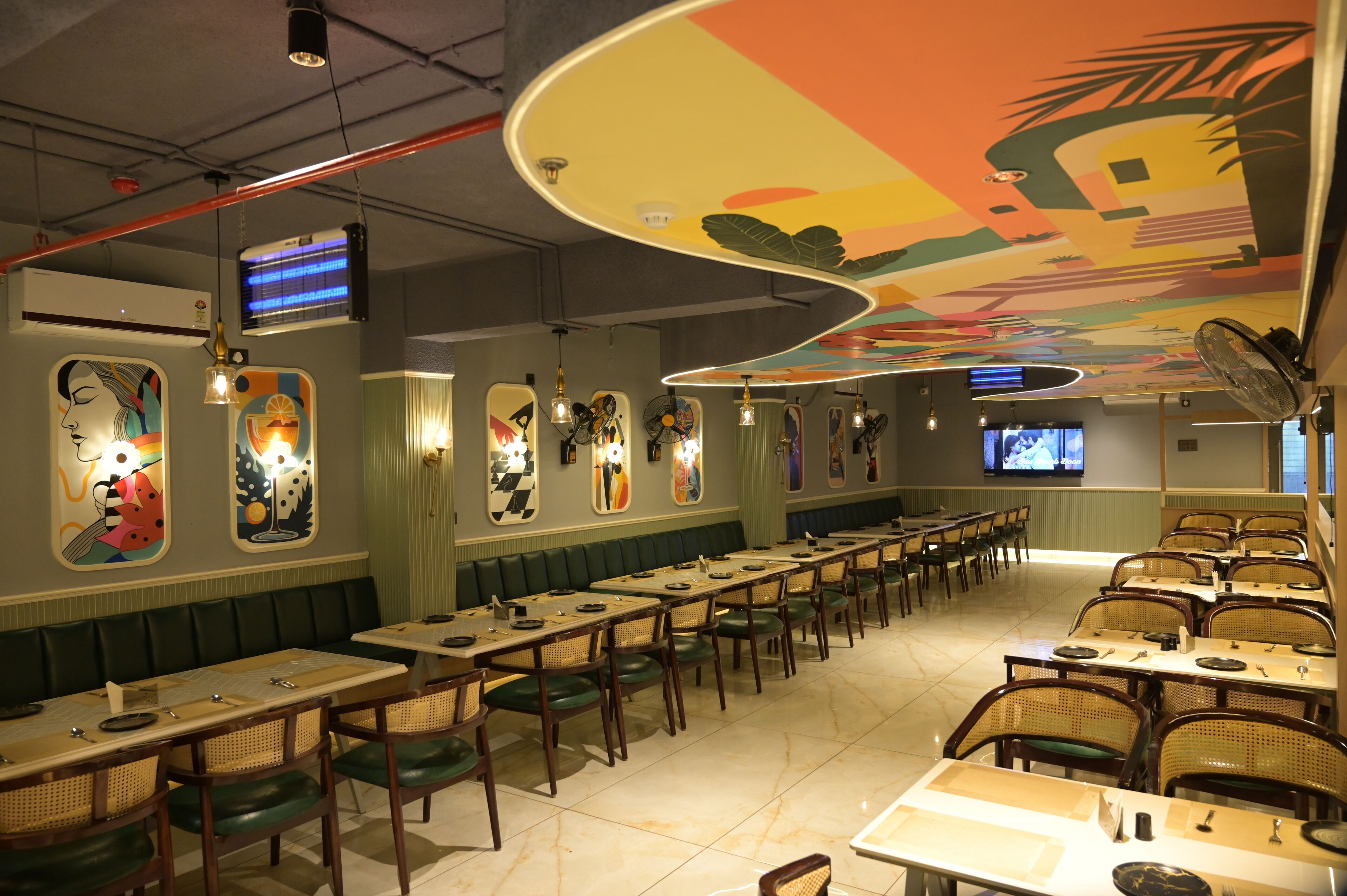 Un restaurant doté de longues tables communes et de banquettes, d’une fresque colorée au plafond et d’un écran de télévision fixé au mur.