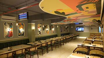 Un restaurant doté de longues tables communes et de banquettes, d’une fresque colorée au plafond et d’un écran de télévision fixé au mur.