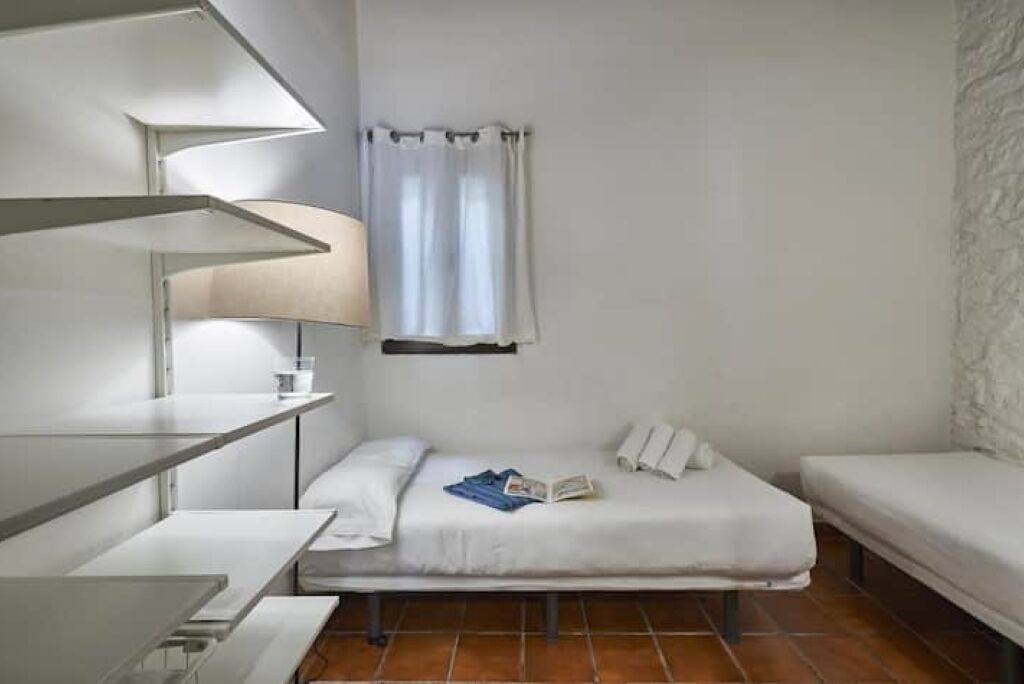 A small, modern room with a single bed, a bedside table, a lamp, and a window with curtains.