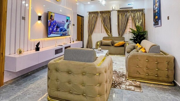A modern living room with a flat-screen TV, a white console table, two tufted sofas, a coffee table, and a patterned rug. - La Veda Manor Luxury Apartment (Ibadan)