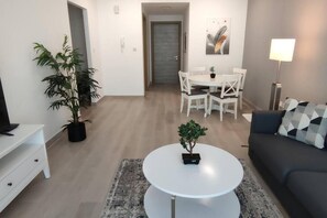 Ein modernes Wohnzimmer mit einem weißen runden Couchtisch, einem grauen Sofa, einem Essbereich mit weißen Stühlen, einer Topfpflanze und einer Lampe.