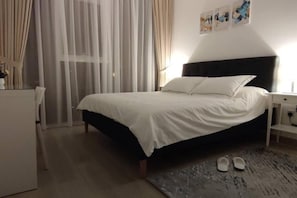 Ein Schlafzimmer mit einem Bett, einem Nachttisch, einem Schreibtisch, einem Sessel und einem Paar Hausschuhen auf dem Boden.
