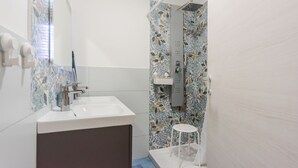 A bathroom with a white sink, a mirror, a shower with floral tiles, and a small white stool. - Pino Baroque near Beach & Etna by Wonderful Italy (Catania)
