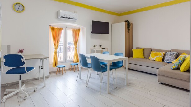 A modern living room with a dining area, a sofa with colorful cushions, a flat-screen TV, and a clock on the wall. - Pino Baroque near Beach & Etna by Wonderful Italy (Catania)