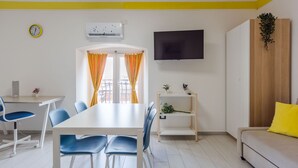 A modern living room with a dining area, a flat-screen TV mounted on the wall, and a wooden plant shelf. - Pino Baroque near Beach & Etna by Wonderful Italy (Catania)