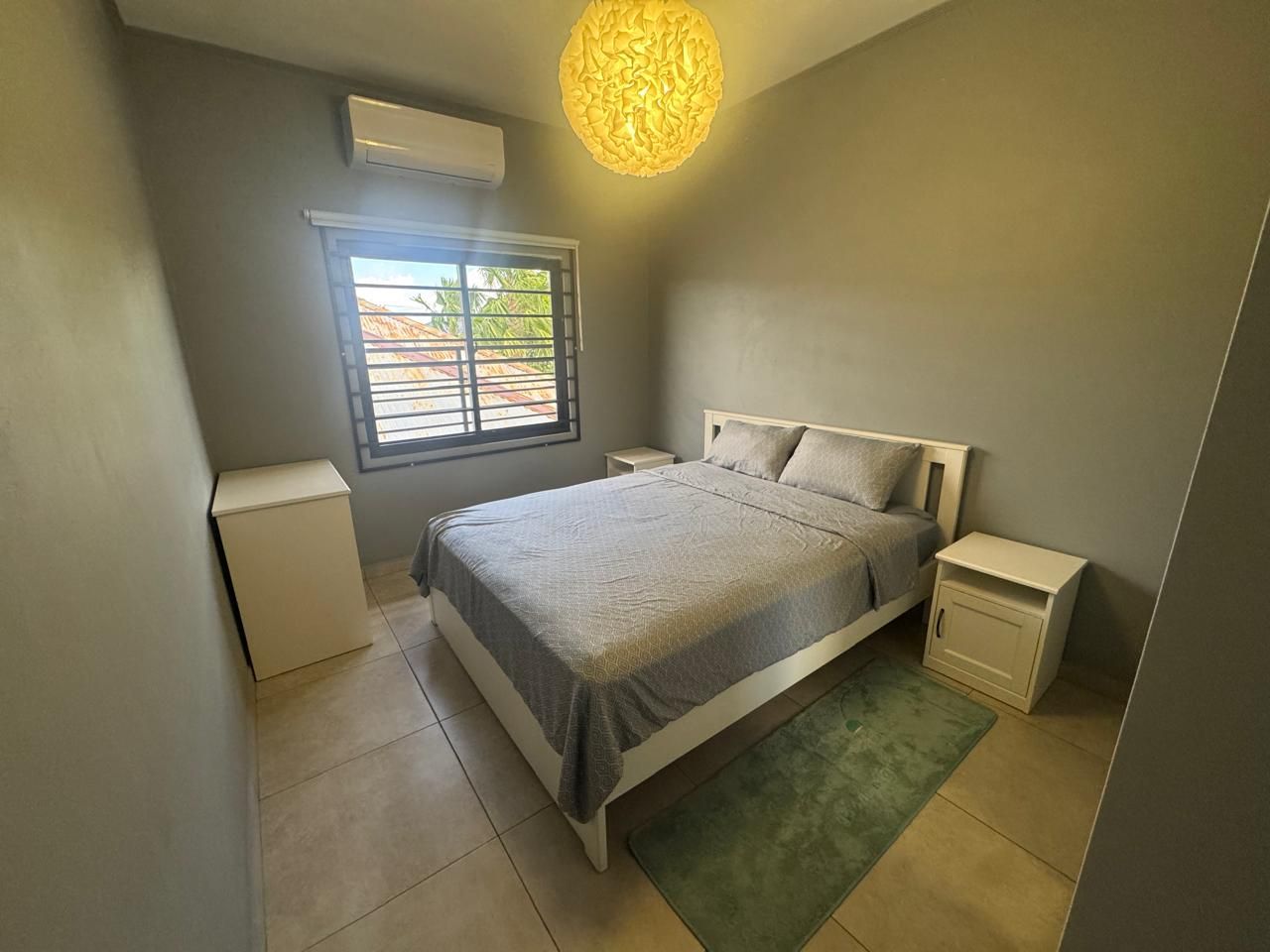A bedroom with a bed, bedside tables, a nightstand, a window, and an air conditioning unit.