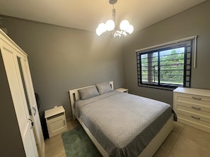 Een slaapkamer met een bed, nachtkastjes, een dressoir, een raam met jaloezieën en een plafondlamp.