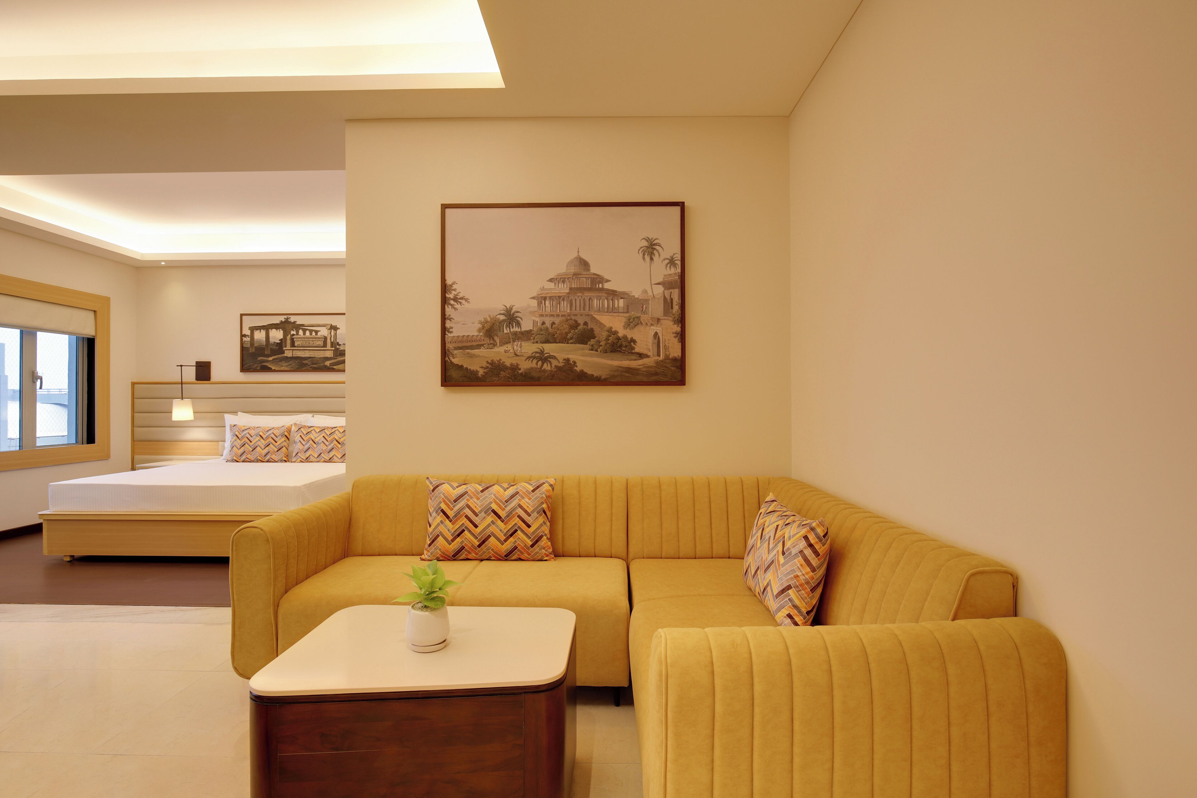 A modern hotel room with a yellow sofa, a bed with a wooden headboard, and a large framed picture of a historical building on the wall.