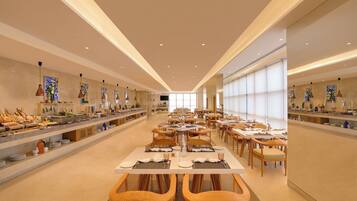 Un grand espace dédié à la salle à manger, avec plusieurs tables et chaises, un buffet situé à gauche et de grandes fenêtres à droite.