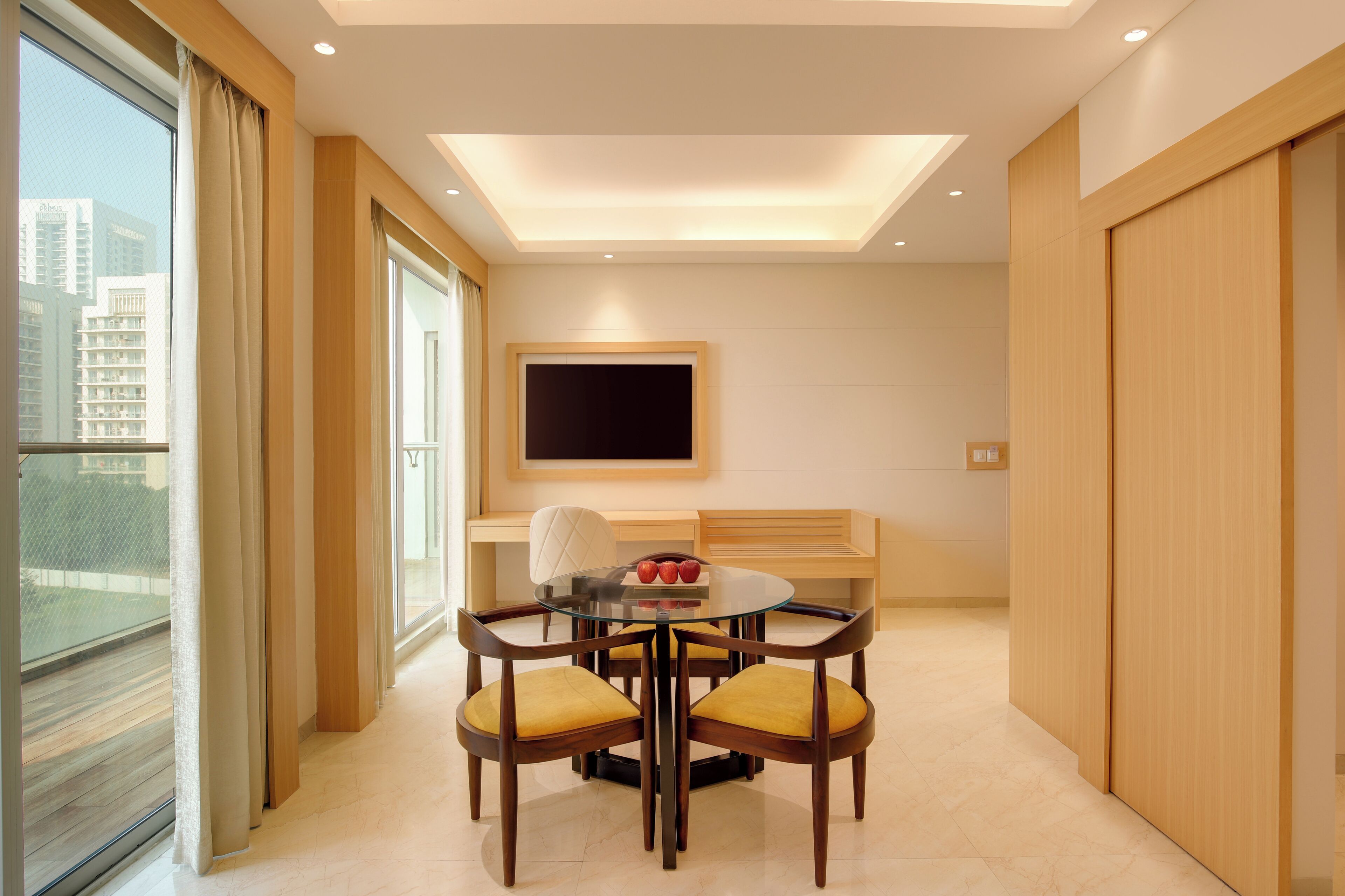 A modern hotel room with a glass dining table, wooden chairs, a flat-screen TV mounted on the wall, and a view of the city through large windows.