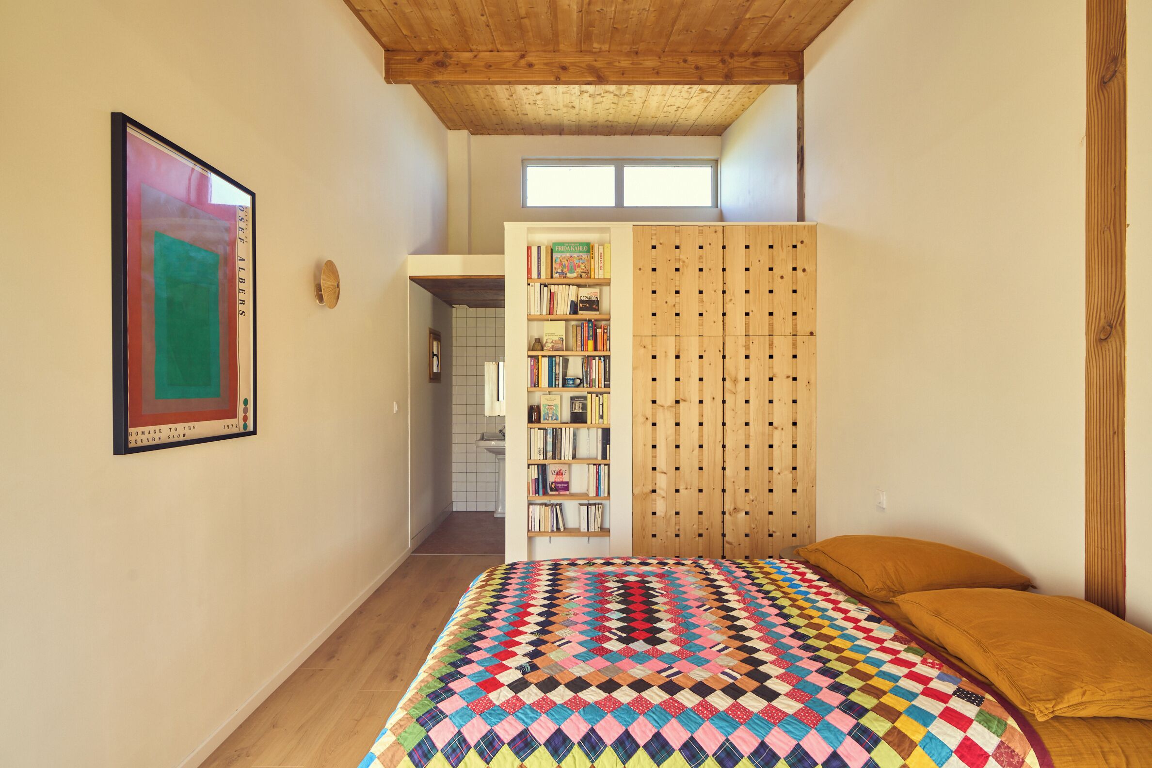 A bedroom with a colorful quilt, a bookshelf, and a wooden wardrobe.
