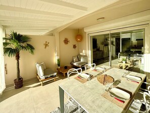A spacious dining area with a large table set for four, surrounded by white chairs. There is a small round table with a single chair, and a cushioned bench with a pillow. A potted plant is visible on the left side. - 2-bedroom condo in soothing Orient Bay. (Orient bay)