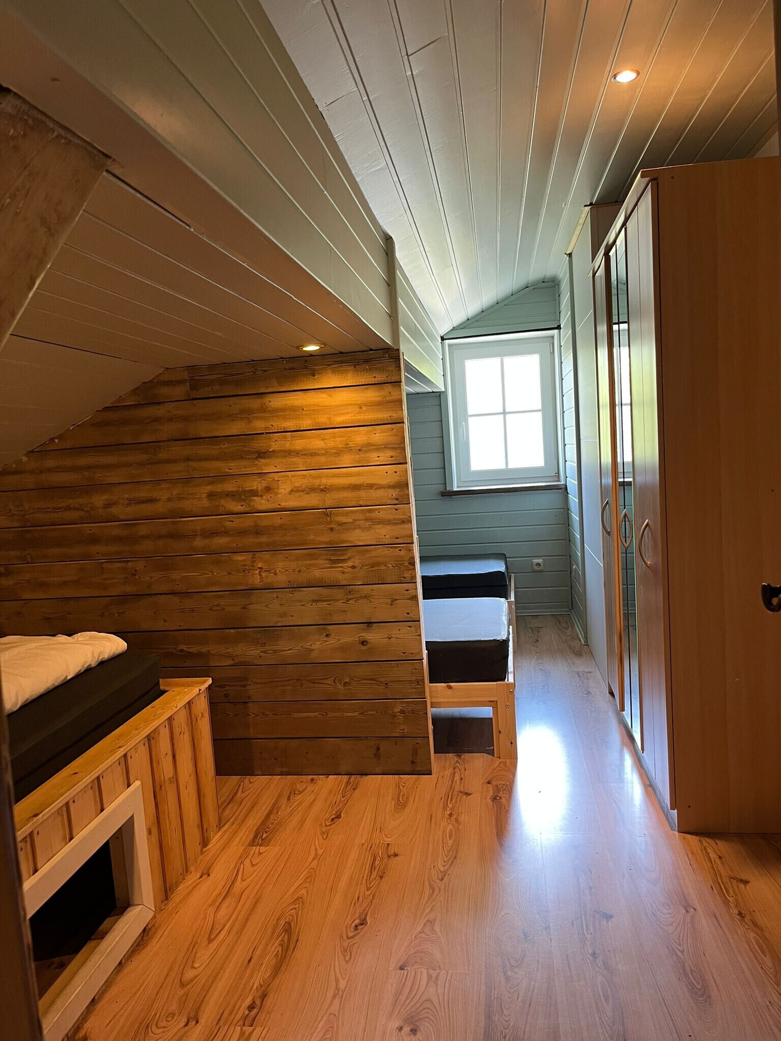 A wooden cabin interior with a loft bedroom, a small window, and a wooden wardrobe.