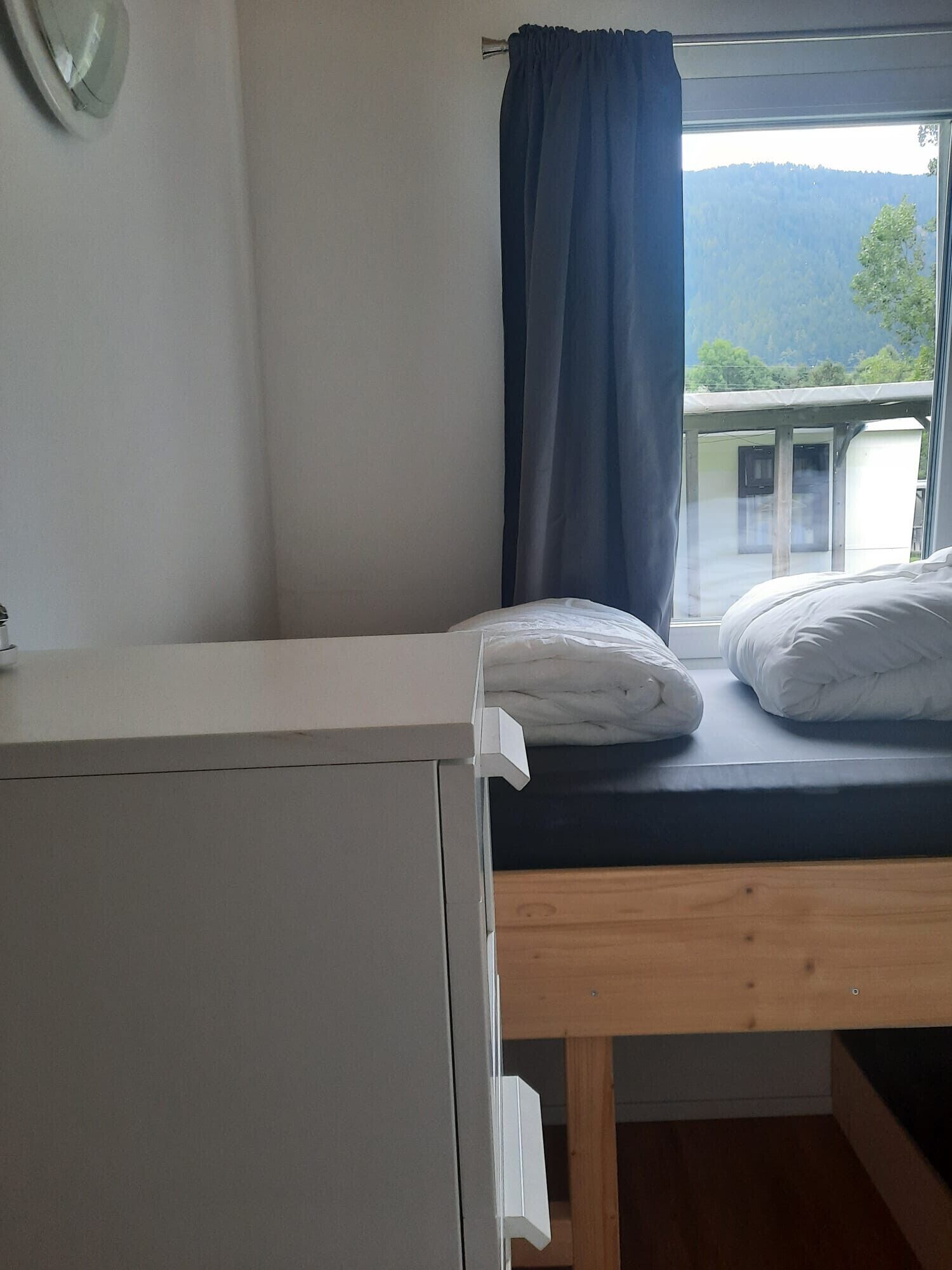 A single bed with a dark mattress and white pillows, a wooden headboard, and a window with a view of a mountainous landscape.