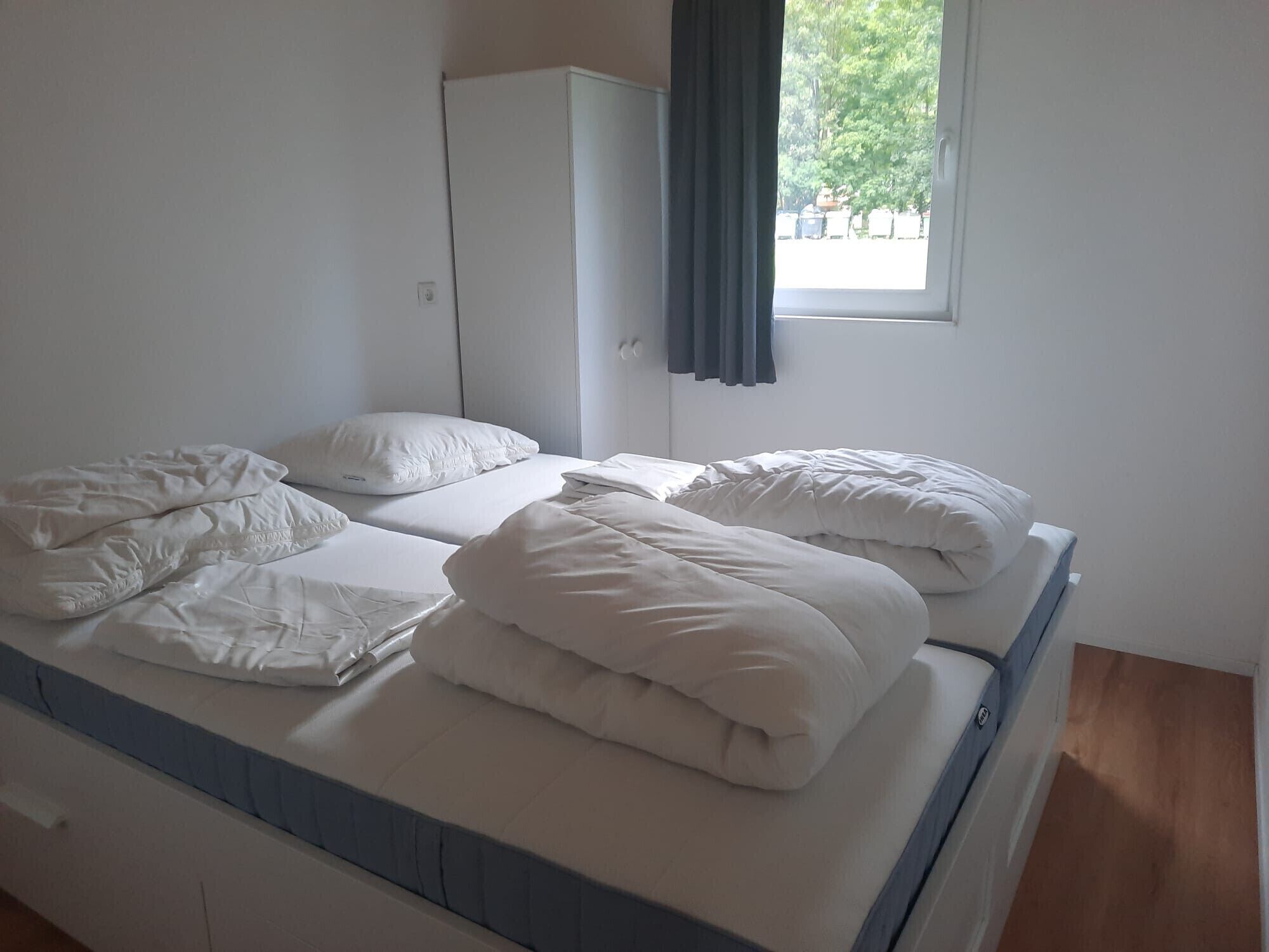 A single bed with white bedding and a blue headboard in a room with a wardrobe and a window with curtains.