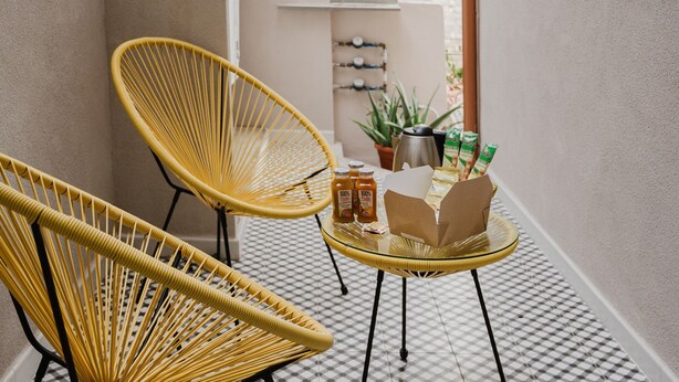 Un petit espace extérieur clos, comprenant deux fauteuils en osier jaune, une table en verre avec une boîte et des bouteilles marron, et une plante en pot.