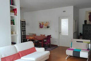 Ein Wohnzimmer mit einem weißen Sofa, einem farbenfrohen Sessel, einem Esstisch mit Stühlen, einem Bücherregal und einer Tür.