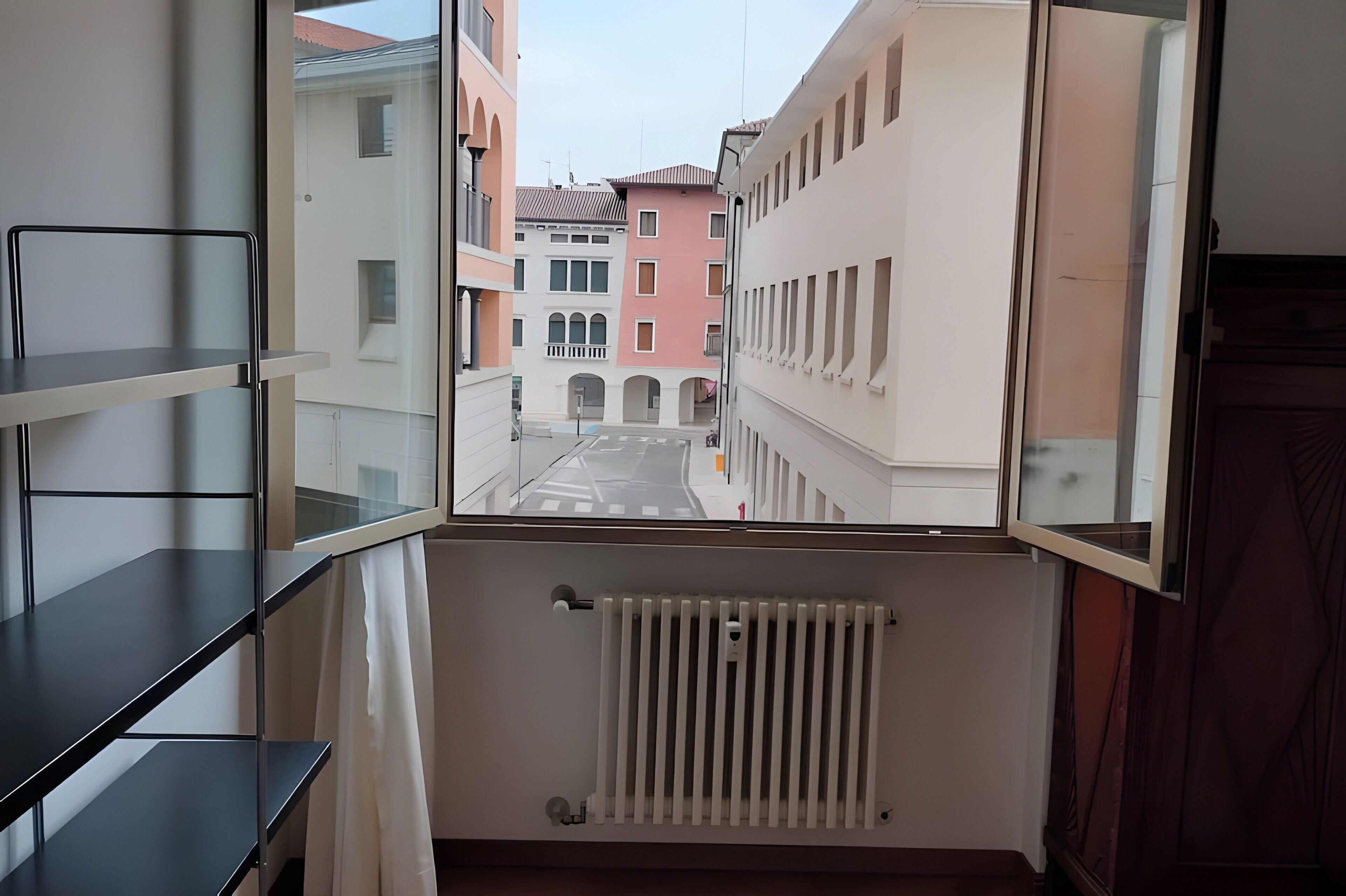 Une vue depuis un balcon, avec un radiateur blanc, une chaise en métal noir et un aperçu des bâtiments voisins.