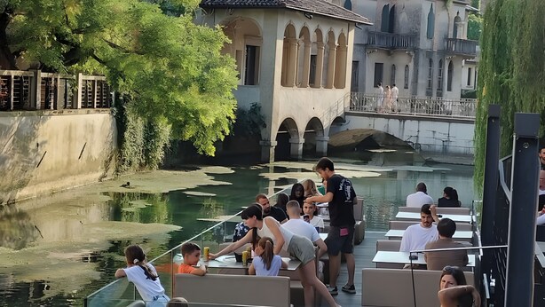 Un restaurante a la orilla del río con terraza, un puente y un edificio con arcos.