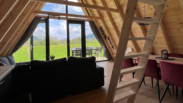 A cozy attic room with a wooden ceiling, a black sofa, a dining table with chairs, and a view of a green field. - Parc Sonnleiten - Edelhert (Gnesau)