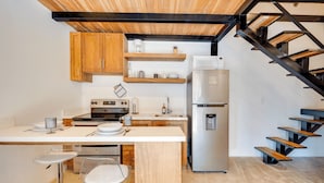 A modern kitchen with wooden cabinets, a stainless steel refrigerator, a built-in oven, and a bar area with stools. - Playa Grande Apartment Loft # 1 - Starlink WiFi - Walk to Beach - Sleeps 3 (Santa Cruz (Playa Grande is a beach community within the Santa Cruz canton))
