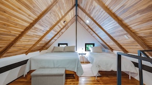 A cozy attic bedroom with wooden beams, two beds, a bench, and a view of greenery outside. - Playa Grande Apartment Loft # 1 - Starlink WiFi - Walk to Beach - Sleeps 3 (Santa Cruz (Playa Grande is a beach community within the Santa Cruz canton))