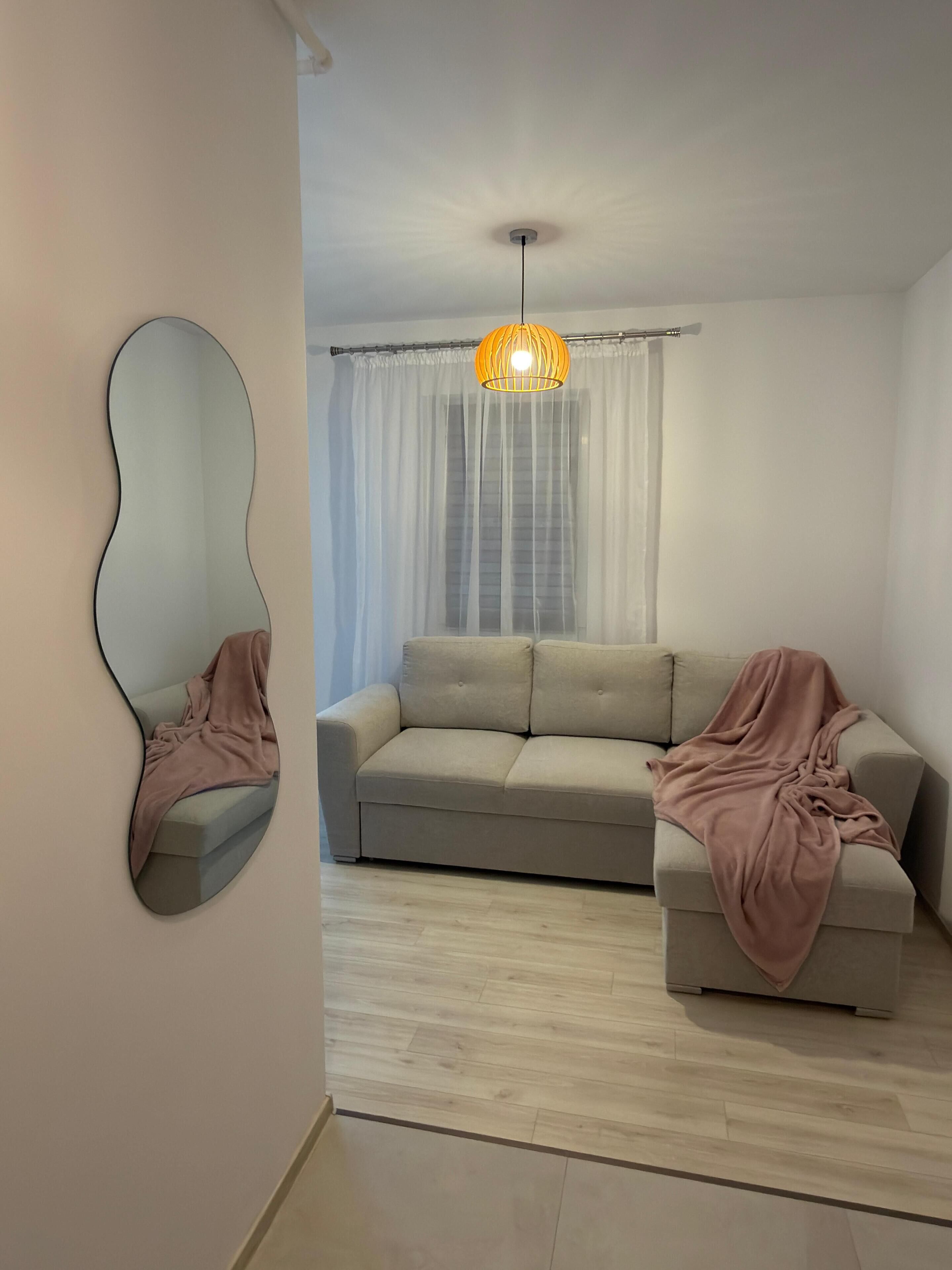 Ein Wohnzimmer mit einer Couch, einem großen Spiegel, einer Pendelleuchte und einem Fenster mit transparenten Vorhängen.