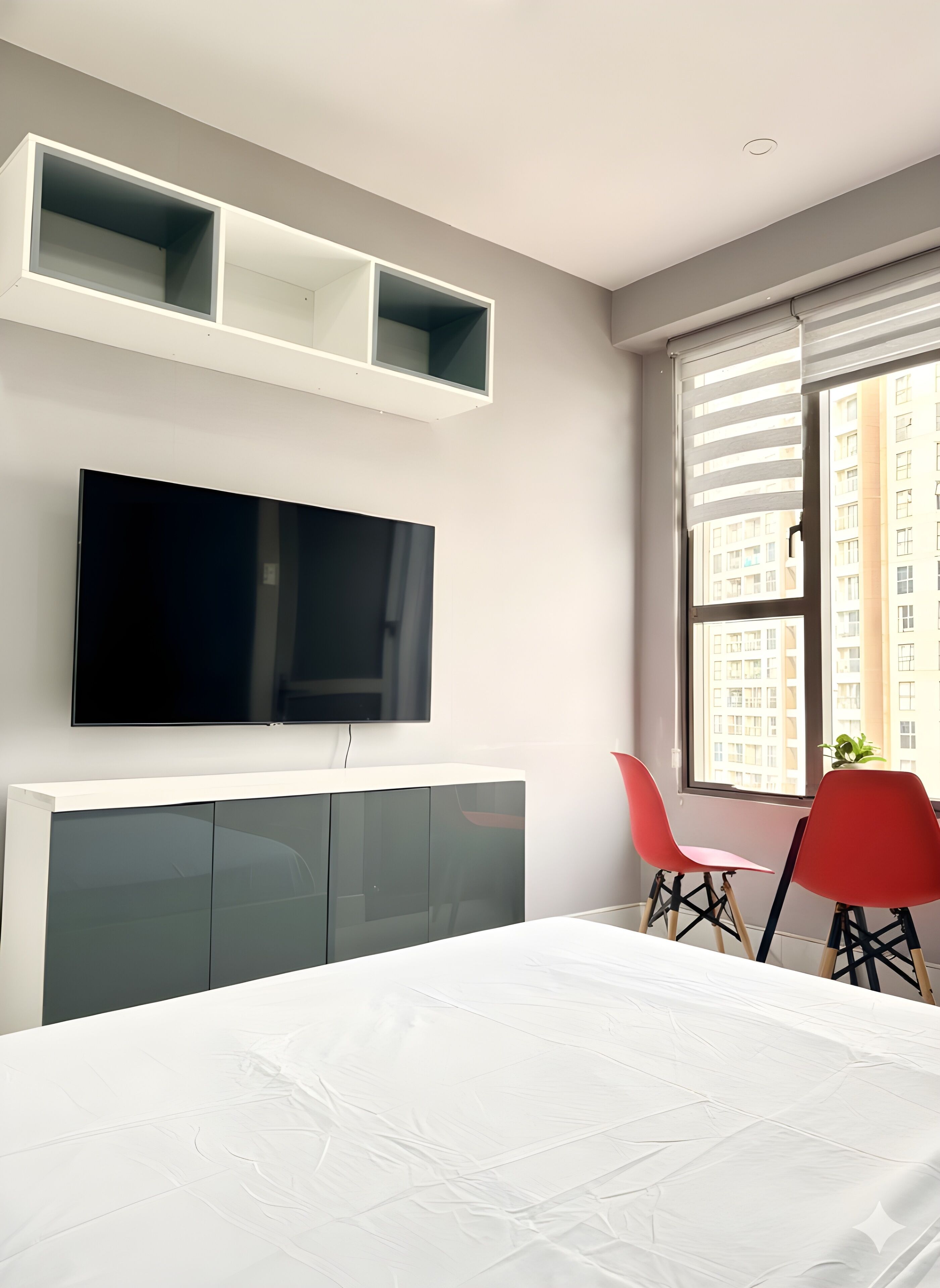 A modern hotel room with a large bed, a flat-screen TV mounted on the wall, a small table with two red chairs, and a window with a view of buildings.